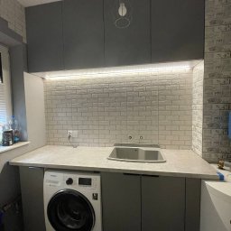 Firma meblowa Anzhela Dzhvarelia - Szara zabudowa kuchenna z pralką pod blatem, biała ceglana ściana z oświetleniem LED podszafkowym i minimalistyczną lampą wiszącą.