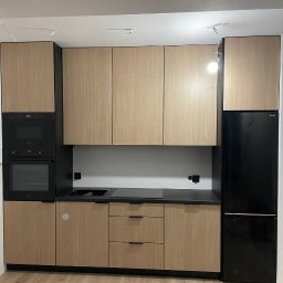 Firma meblowa Anzhela Dzhvarelia - Nowoczesna zabudowa kuchenna z drewnopodobnymi frontami, czarnym blatem i sprzętem AGD w czarnych obudowach, widoczne niedokończone podłączenie oświetlenia sufitowego.