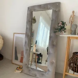 Pełnowymiarowe lustro w ramie obitej szarym, pluszowym materiałem z guzikami, ustawione w jasnym pokoju z minimalistycznym wystrojem, obok obrazu w ramie i książek.