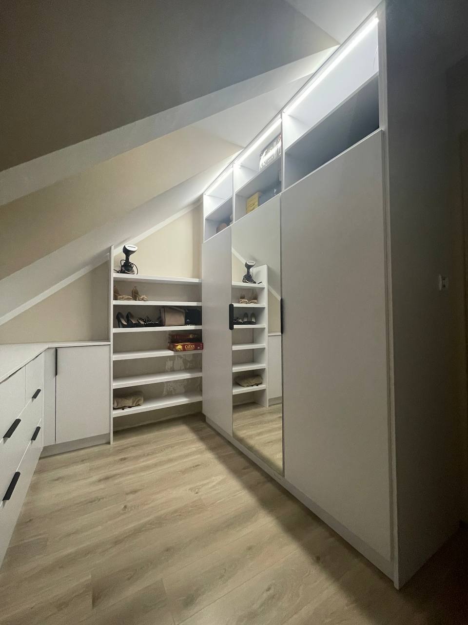 Biała garderoba w skosach poddasza z oświetleniem LED, półkami na buty i lustrem na drzwiach przesuwnych, widoczna gra Catan na półce.