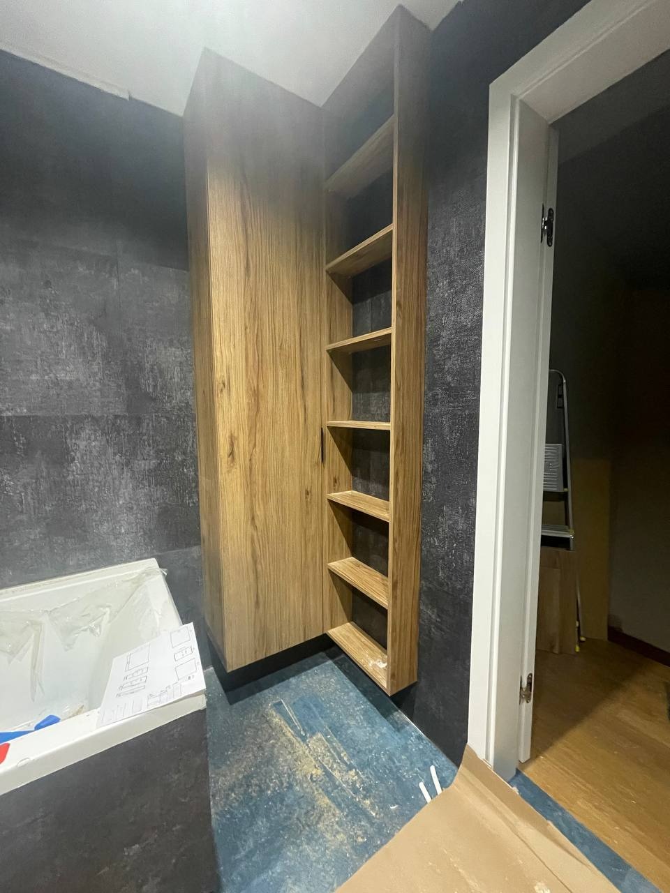 Wysoka, drewniana szafa z półkami w surowym, szaro-niebieskim wnętrzu łazienki w trakcie remontu, widoczna wanna z folią ochronną i drabina w tle.