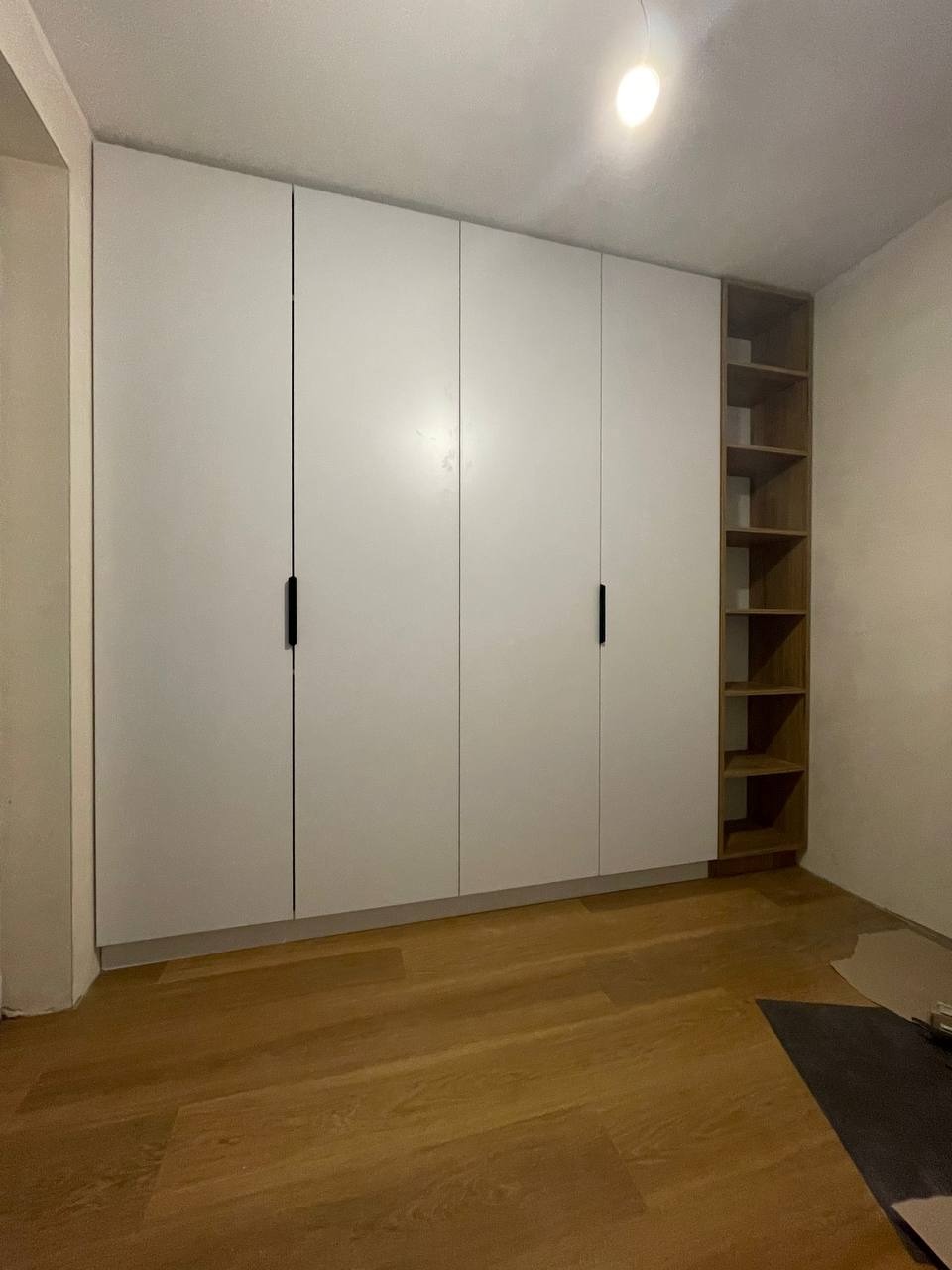 Biała szafa z czterema drzwiami z czarnymi, pionowymi uchwytami, z otwartymi półkami po prawej stronie, na podłodze panele w kolorze drewna