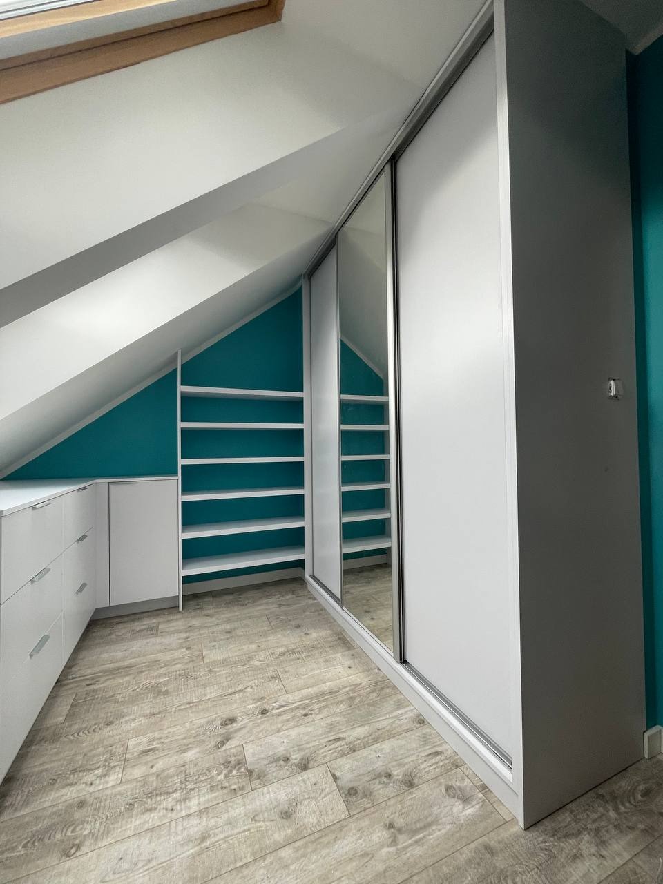 Biała szafa z przesuwnymi drzwiami i lustrem, dopasowana do skosu poddasza, z otwartymi półkami po lewej stronie, na podłodze panele imitujące drewno, turkusowa ściana w tle.