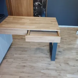 Biurko z blatem z jasnego drewna dębowego, z wysuwaną szufladą na metalowej nodze, na podłodze z paneli drewnianych.