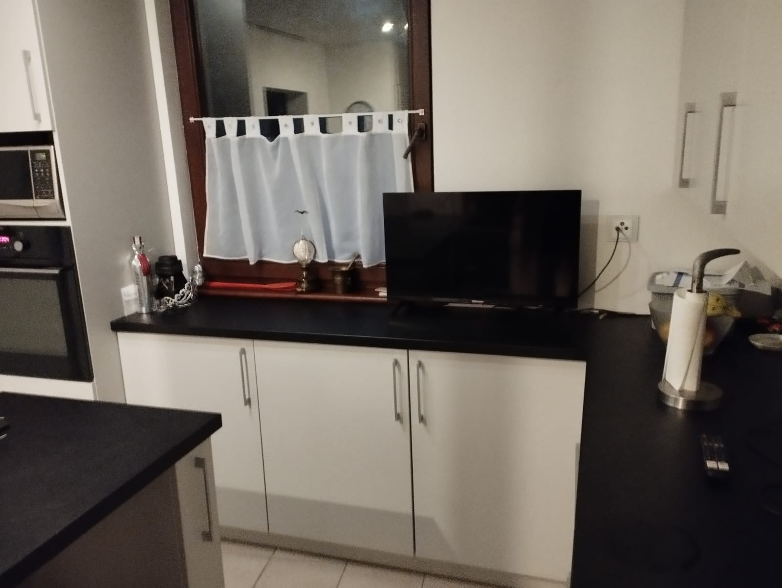 Białe szafki kuchenne pod blatem w zabudowie, z telewizorem i zasłonką w oknie.