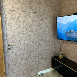 R-PRO - Pokój z ukrytymi drzwiami pokrytymi tapetą w odcieniach beżu i brązu, dopasowaną do wzoru na ścianach, obok stoi czarna szafka RTV z kwiatkiem w złotej doniczce, a nad nią wisi telewizor.