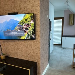 R-PRO - Pokój dzienny z telewizorem wyświetlającym komunikat o braku sygnału, ściana pokryta tapetą o teksturze kamienia, widoczna część kuchni z białymi meblami i złotym okapem.