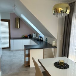 R-PRO - Nowoczesna kuchnia w skosach na poddaszu z białymi szafkami, czarnym blatem, złotym okapem i kulistą lampą nad białym stołem z dekoracją.