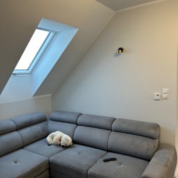 R-PRO - Poddasze z szarą sofą modułową, pluszowy miś na siedzisku, okno dachowe z widokiem na jasne niebo, nowoczesna lampa sufitowa i kinkiet na ścianie.