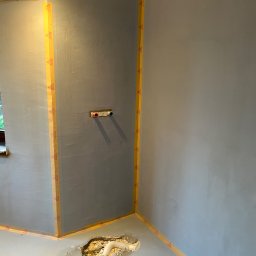 R-PRO - Świeżo nałożona szpachla na ścianach zabezpieczonych taśmą malarską w pomieszczeniu w Olsztynie, widoczne wystające rury instalacji wodnej i otwór w podłodze.