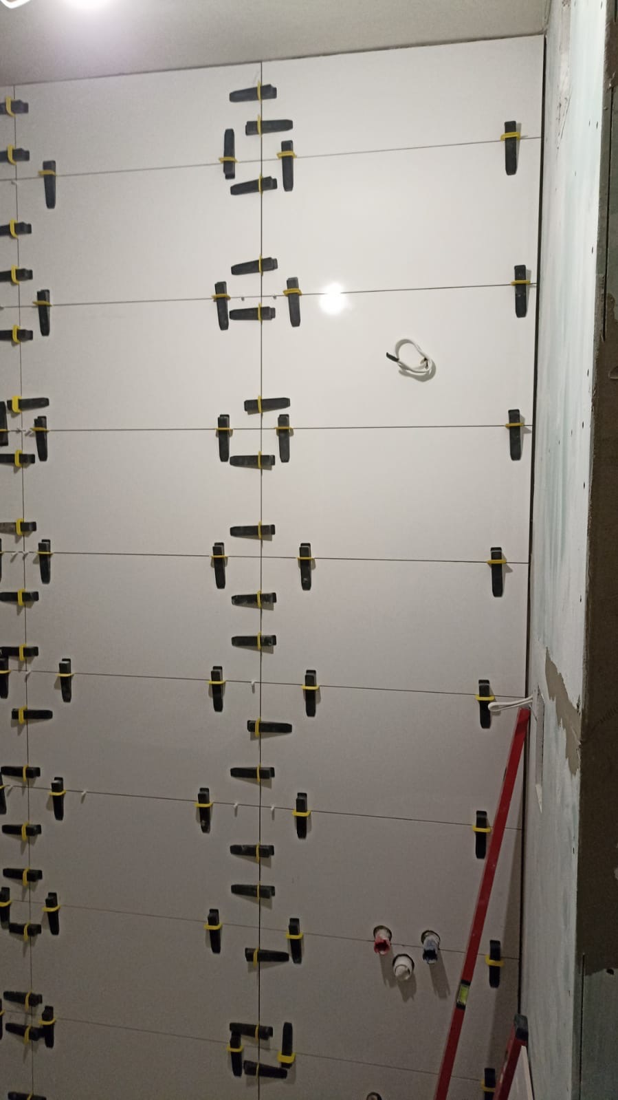 Świeżo ułożone, białe płytki ceramiczne na ścianie z użyciem systemu poziomowania płytek (klipsy i kliny), widoczne przewody elektryczne i fragment poziomicy.