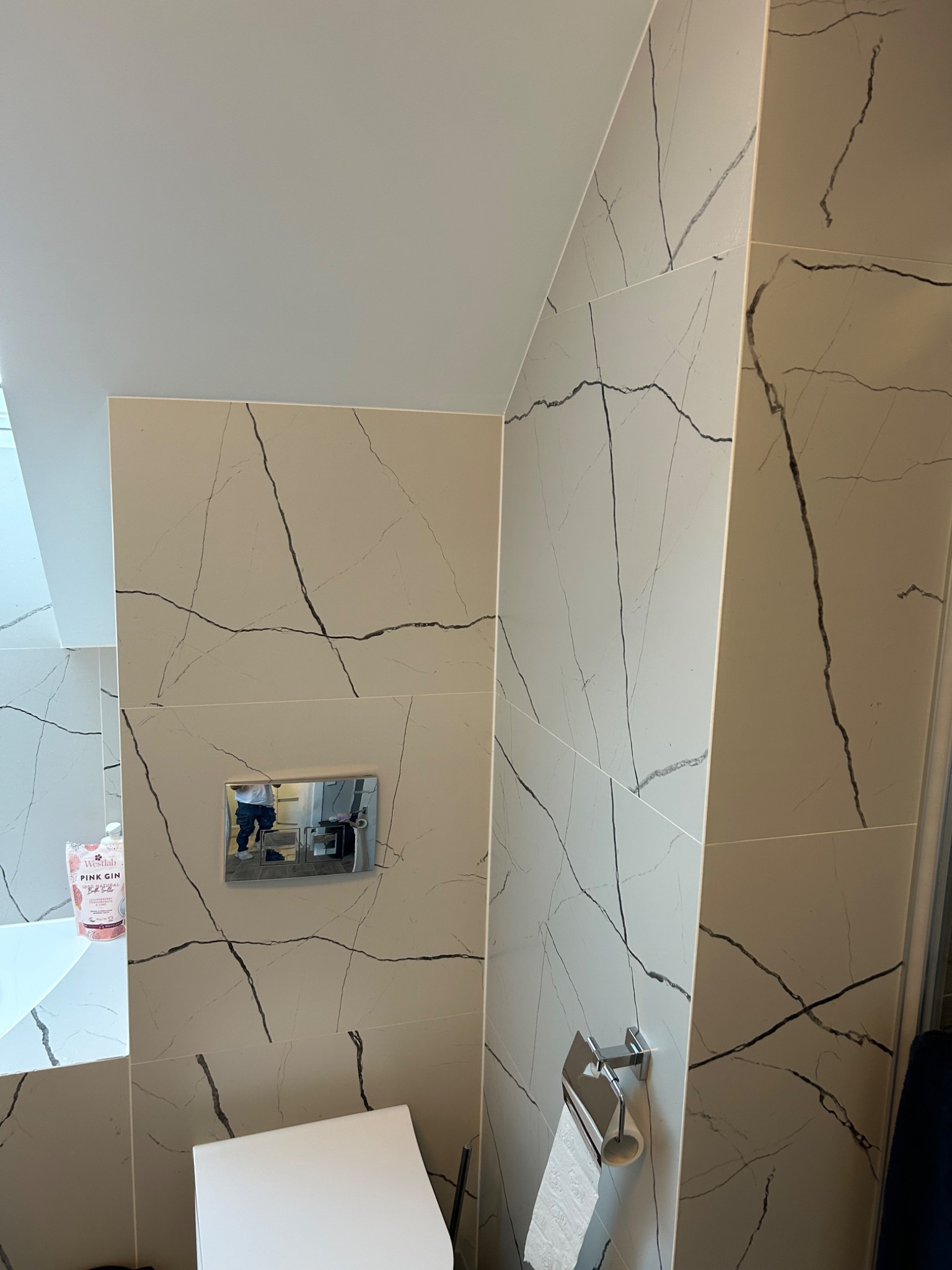 Wykończona łazienka w Olsztynie z płytkami imitującymi marmur z wyraźnym wzorem żyłkowania, wisząca toaleta z chromowanym przyciskiem spłukiwania i uchwytem na papier toaletowy.