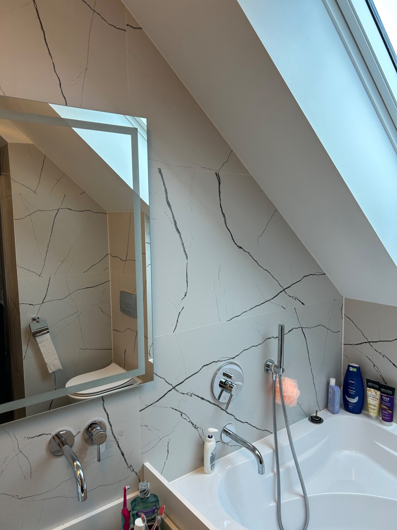 Wykończona łazienka na poddaszu z białą wanną, armaturą w kolorze chromu i płytkami imitującymi marmur z szarymi żyłami, widok na toaletę odbity w lustrze.