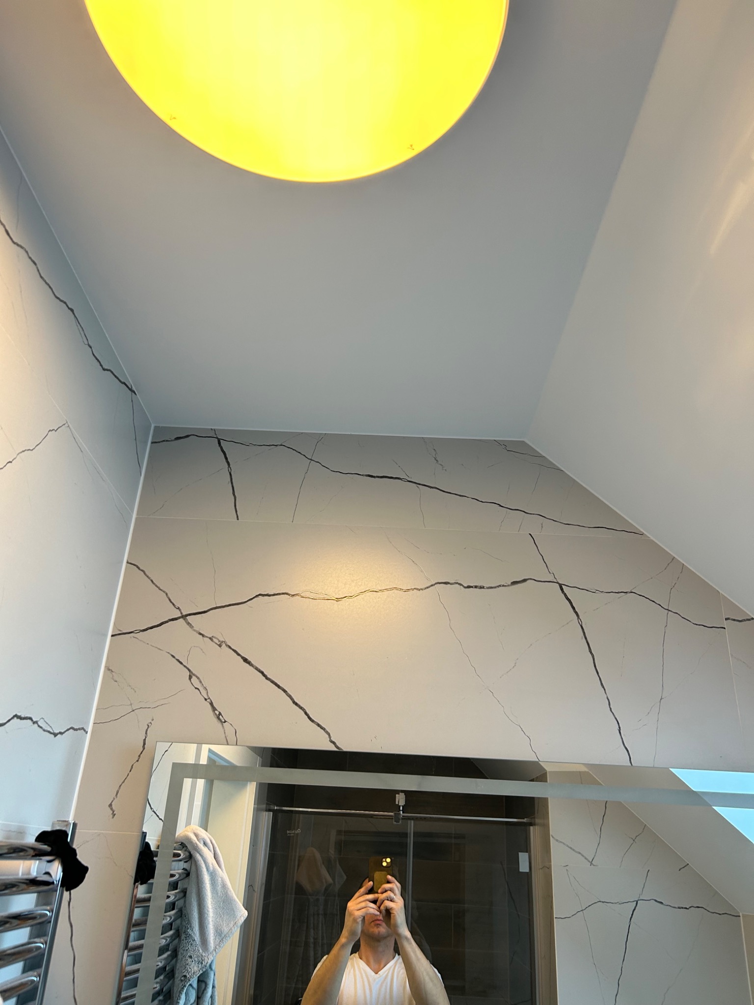 Wykończona łazienka na poddaszu z białymi płytkami imitującymi marmur z czarnymi żyłami, widoczne lustro i oświetlenie sufitowe, odbicie osoby robiącej zdjęcie.