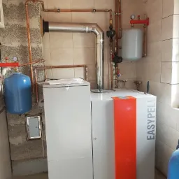 Kotłownia z kotłem na pellet EasyPell, instalacja z miedzianych rur, zbiorniki wyrównawcze w kolorze niebieskim i szarym, płytki ceramiczne na ścianach.