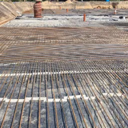 Siatka zbrojeniowa ułożona na folii izolacyjnej w wykopie fundamentowym, widoczne rury instalacyjne i stosy prętów zbrojeniowych w tle.