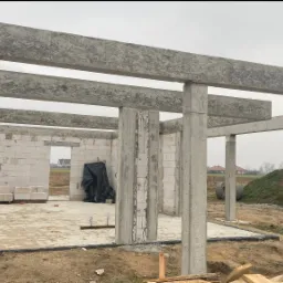 Surowy szkielet domu w budowie z betonu komórkowego i żelbetu, widoczne słupy i belki stropowe, teren budowy z hałdą ziemi w tle.