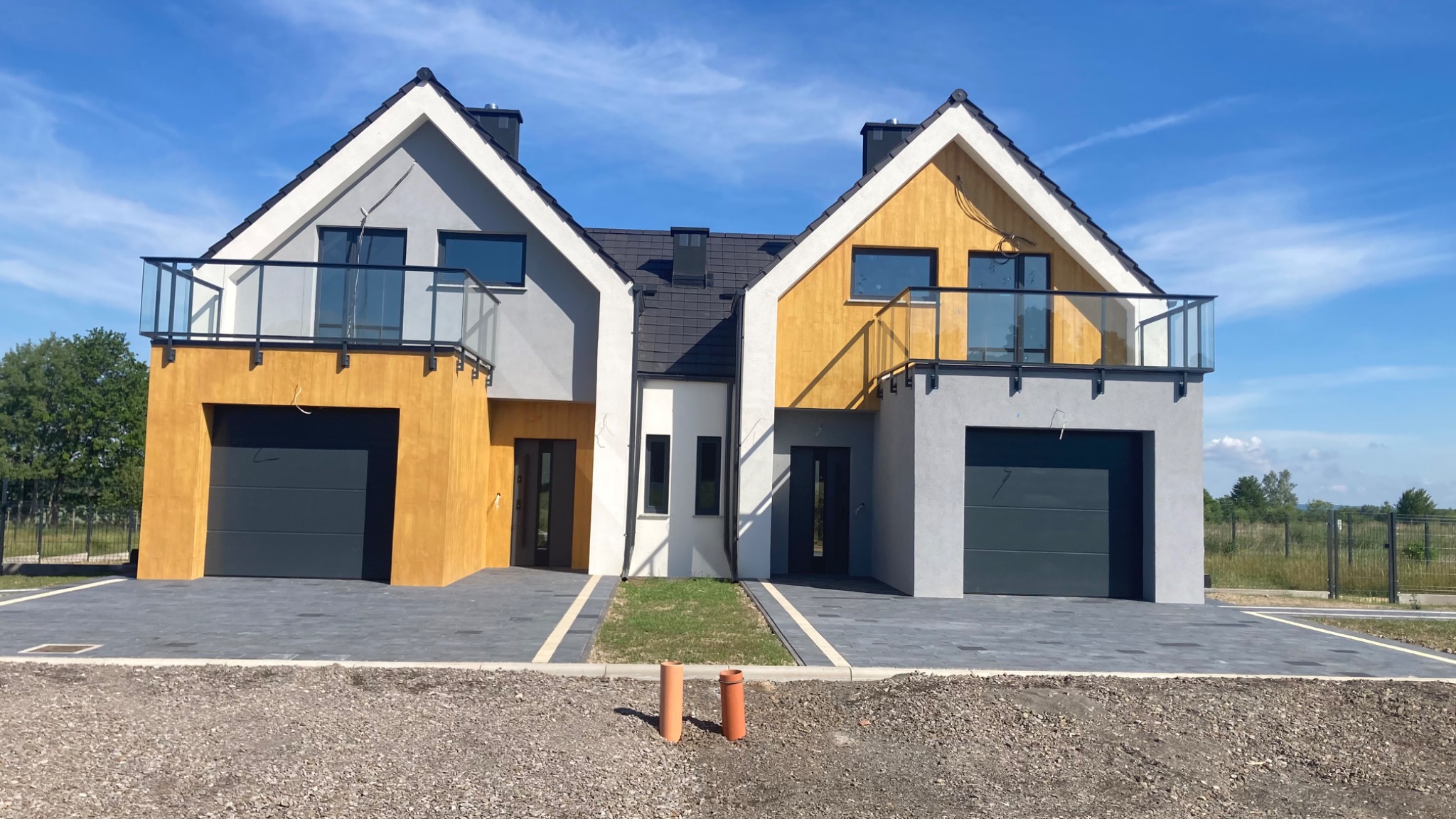 Dwa bliźniacze domy w stanie deweloperskim z balkonami i garażami, wykończone w odcieniach szarości i żółci, z widocznymi elementami instalacji na niezagospodarowanym terenie.