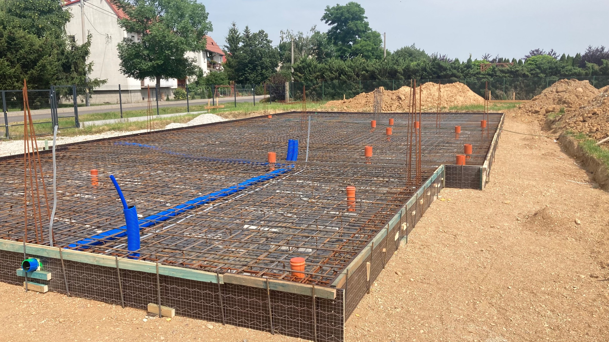 Zbrojenie fundamentów z rurami instalacyjnymi, widoczne szalunki i teren budowy z hałdami ziemi w tle.