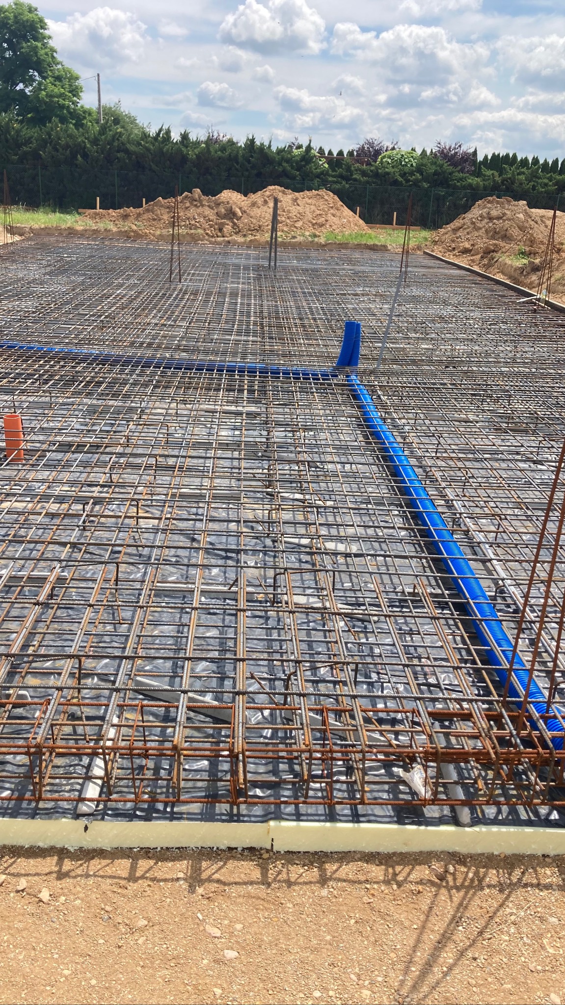 Siatka zbrojeniowa fundamentów ułożona na izolacji termicznej, z widocznymi niebieskimi rurami instalacji podposadzkowej, w tle pryzmy ziemi i zieleń.