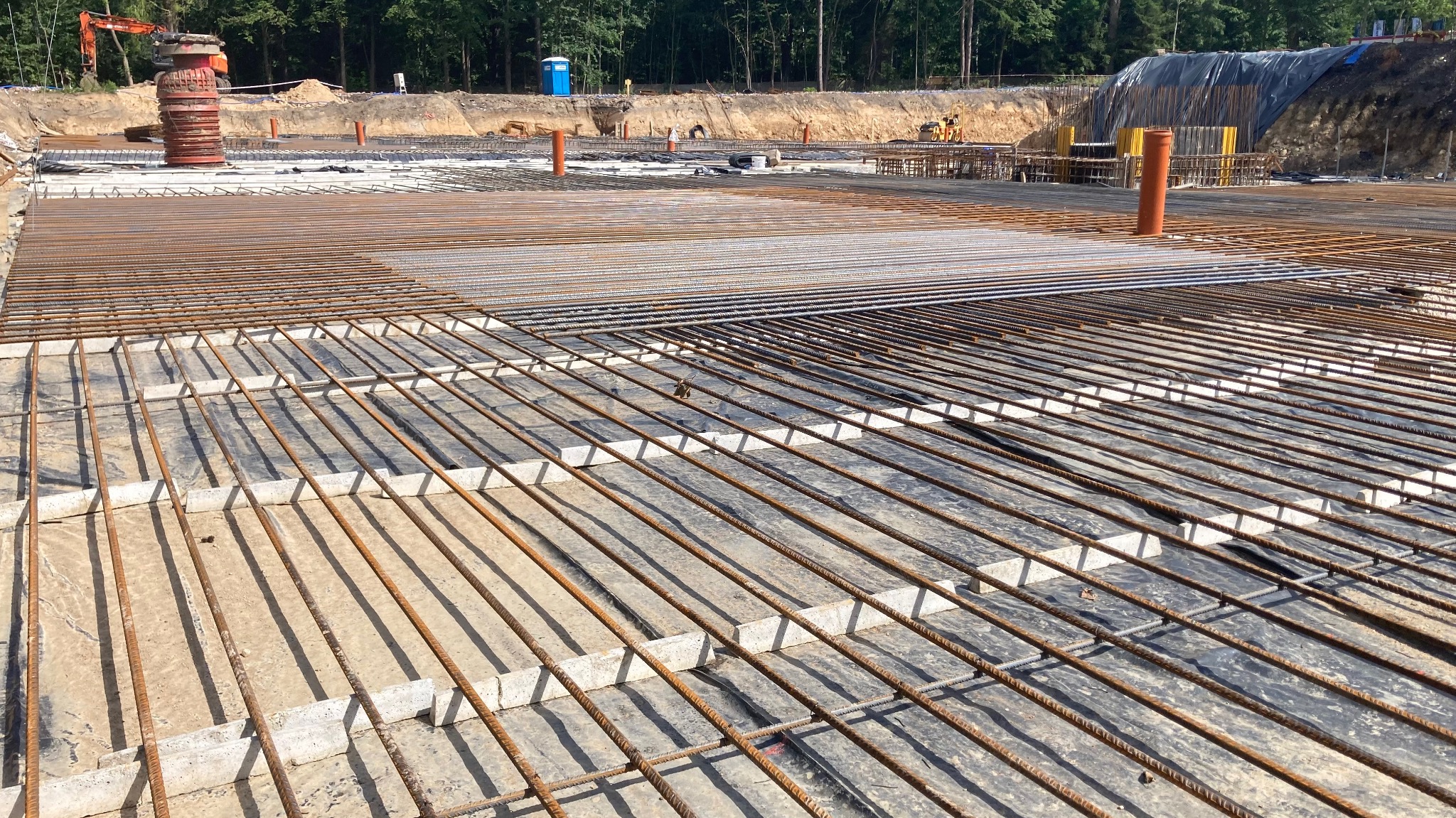 Siatka zbrojeniowa na czarnej folii, podkładki betonowe, w tle wykop z pomarańczowym dźwigiem i przenośna toaleta.