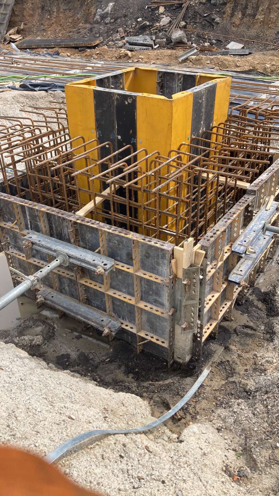 Zbrojenie i szalunek pod fundament z żółtym elementem szalunkowym w kształcie sześcianu, widoczne pręty zbrojeniowe, metalowe płyty szalunkowe i ziemia wokół.