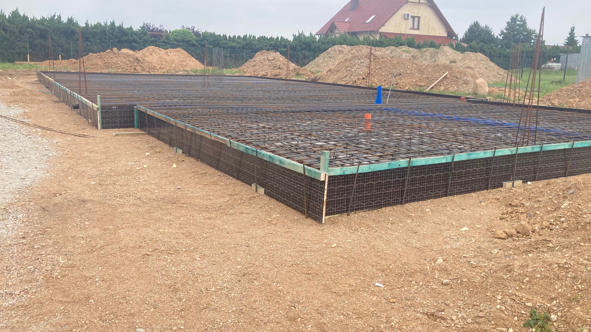 Przygotowany zbrojony szalunek pod płytę fundamentową z widocznymi elementami instalacji wodnej i elektrycznej, w tle hałdy ziemi i dom jednorodzinny.