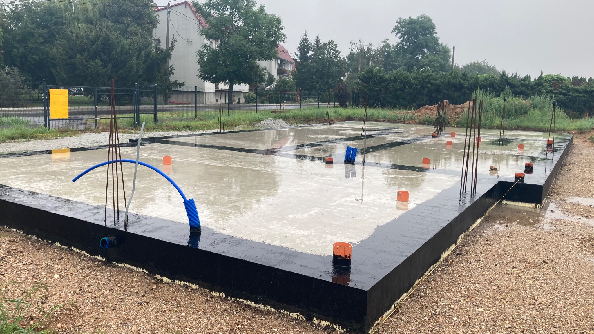 Zalane wodą fundamenty domu jednorodzinnego z wystającymi prętami zbrojeniowymi i pomarańczowymi osłonami, widoczne rury instalacyjne, czarna izolacja pionowa fundamentów.