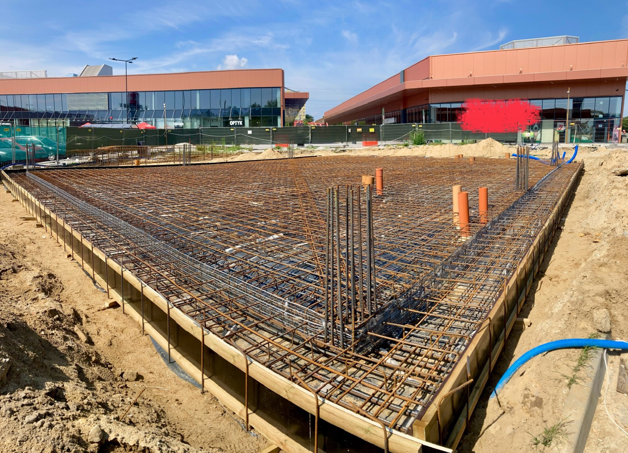 Siatka zbrojeniowa ułożona w szalunku drewnianym, przygotowana do zalania betonem pod fundamenty budynku komercyjnego. Widoczne pionowe pręty zbrojeniowe oraz rury instalacyjne wystające z gruntu.