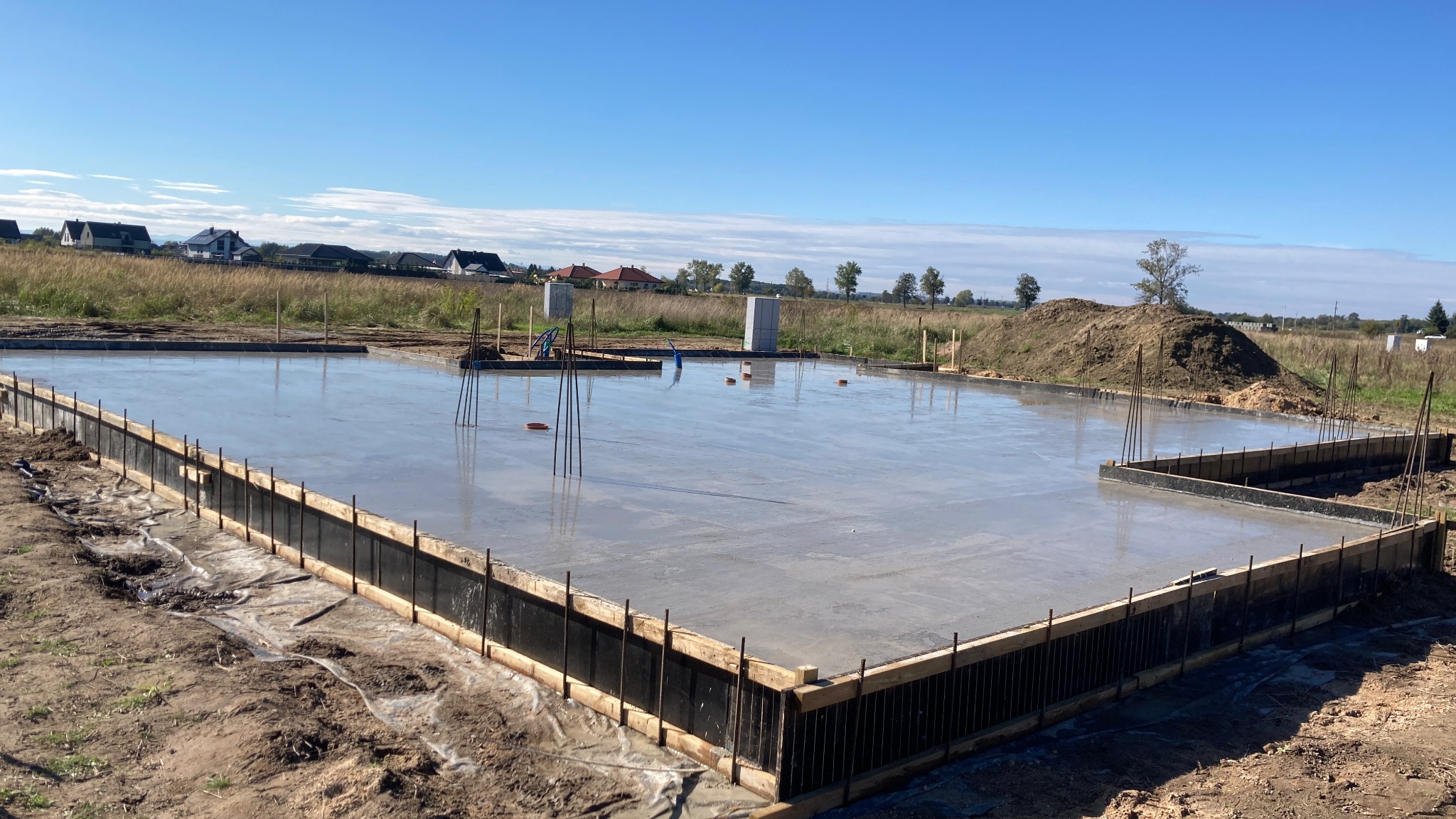 Świeżo wylana płyta fundamentowa z deskowaniem i wystającymi prętami zbrojeniowymi, widok na łąkę i zabudowania w tle, słoneczny dzień.