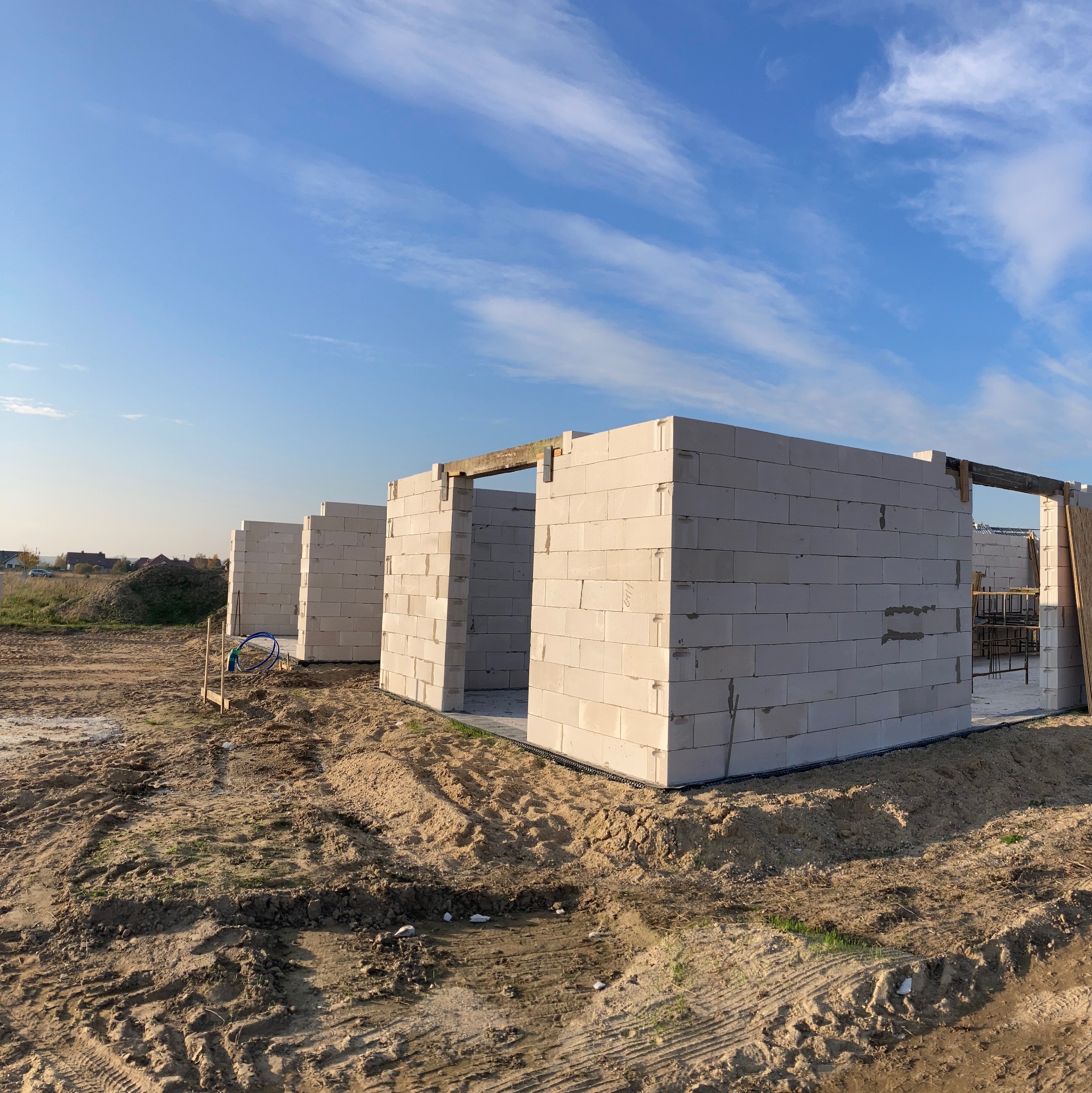 Wznoszenie ścian parteru z betonu komórkowego na fundamencie, widoczne drewniane nadproża i zbrojenie w tle, teren budowy z wykopaną ziemią.