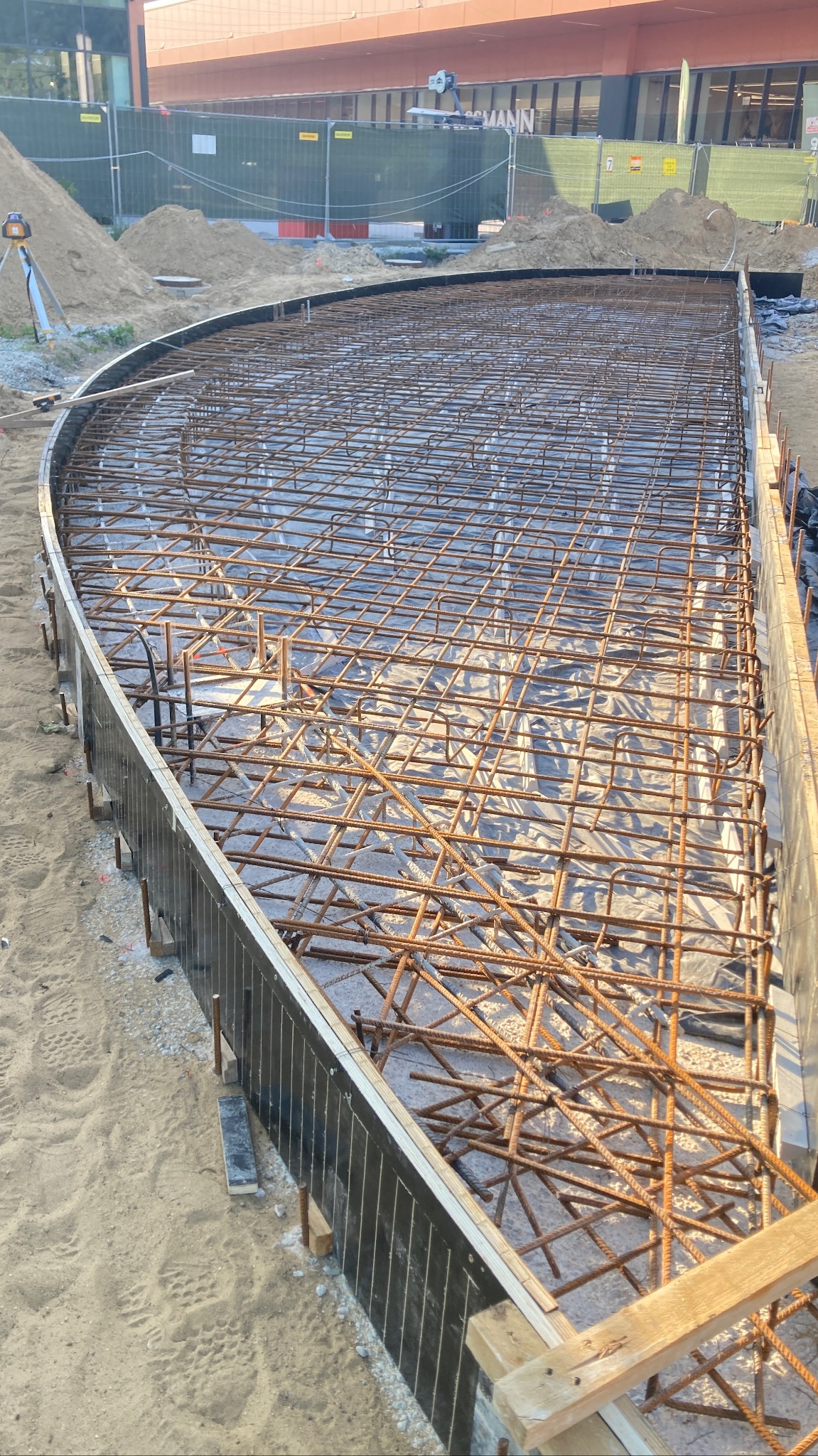 Przygotowanie zbrojenia pod wylewkę fundamentu o nietypowym, łukowatym kształcie, widoczne deskowanie, folia izolacyjna i laserowy niwelator w tle.