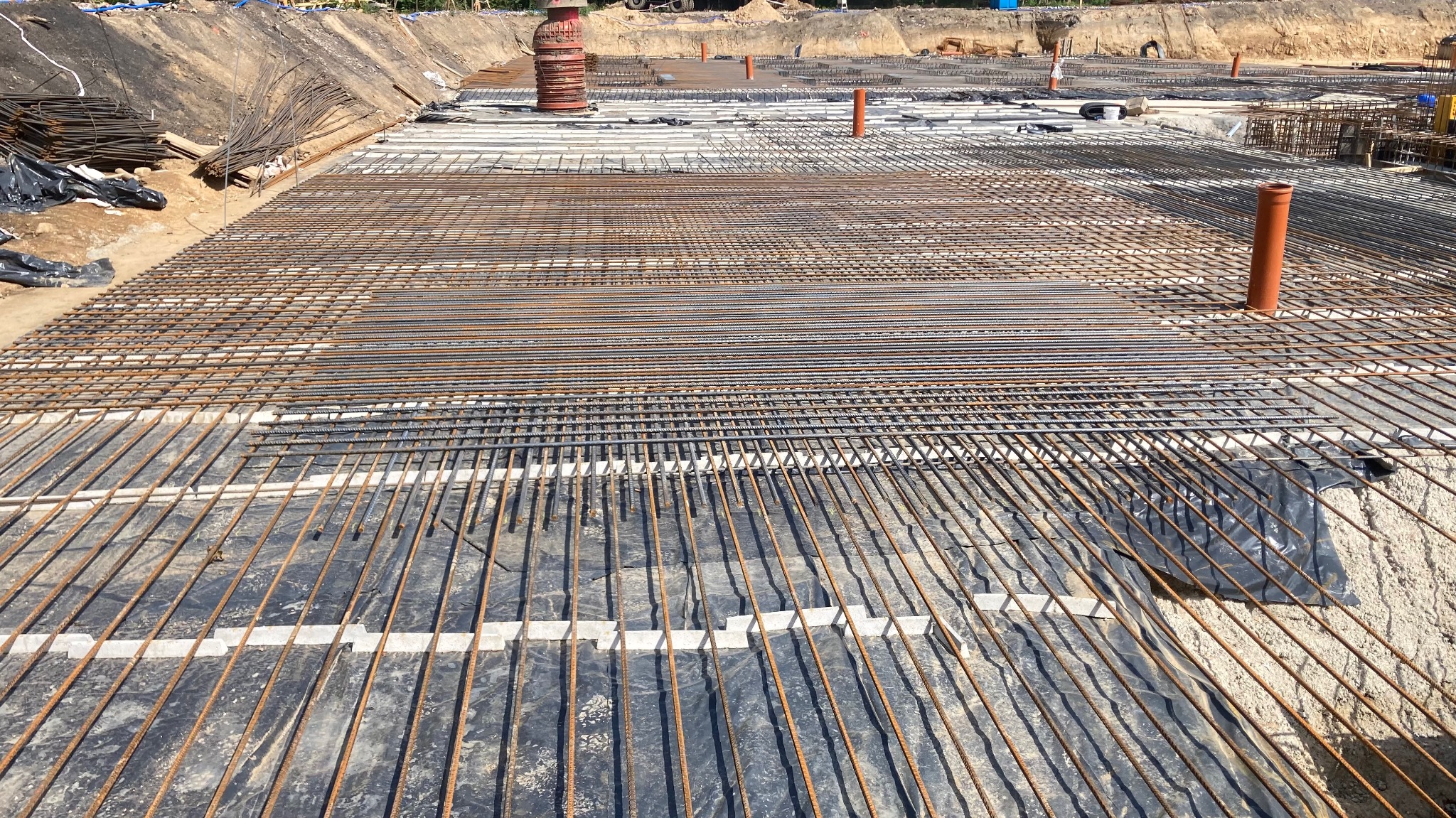 Siatka zbrojeniowa ułożona na folii izolacyjnej w wykopie fundamentowym, widoczne rury instalacyjne i stosy prętów zbrojeniowych w tle.