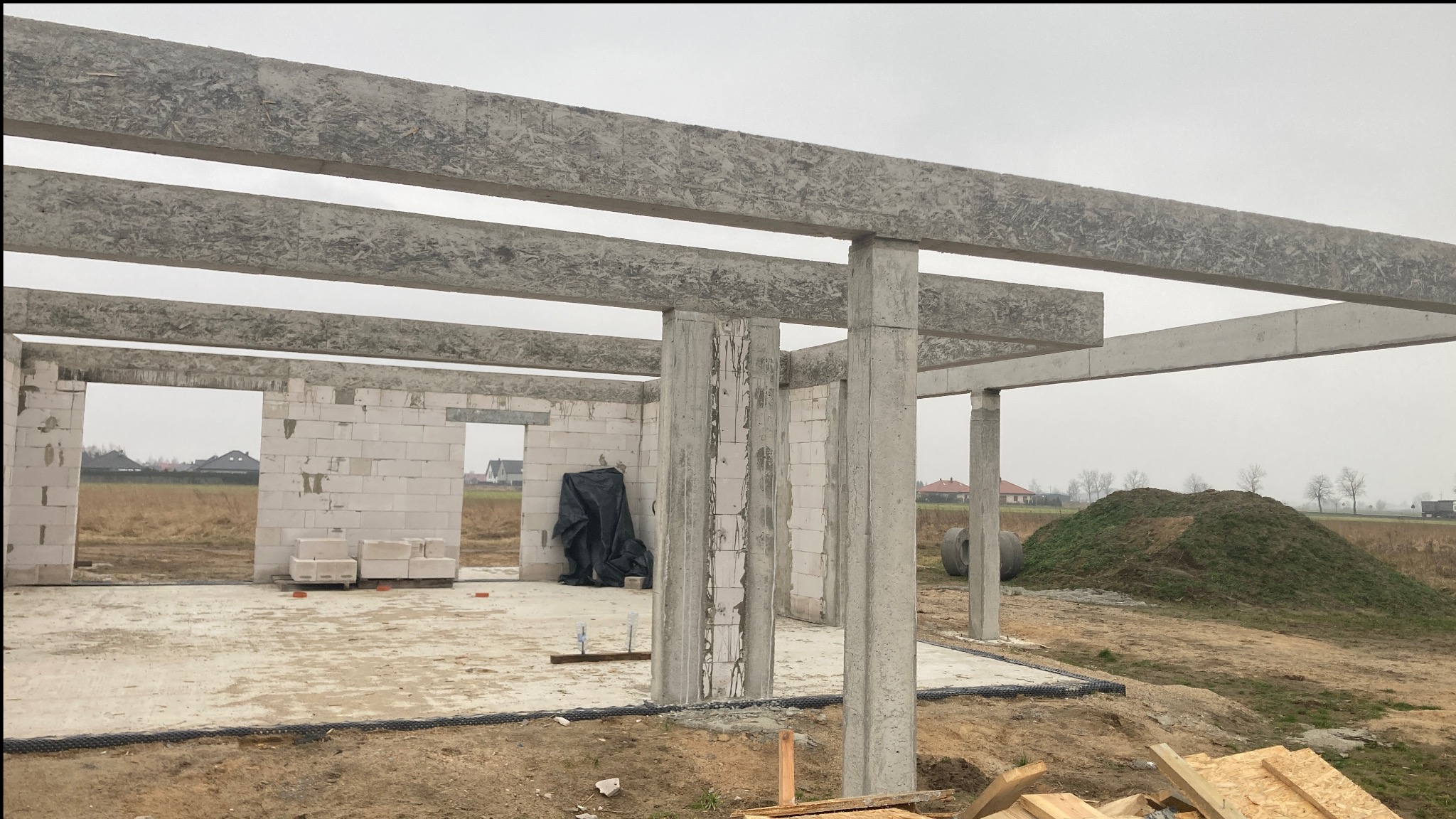 Surowy szkielet domu w budowie z betonu komórkowego i żelbetu, widoczne słupy i belki stropowe, teren budowy z hałdą ziemi w tle.