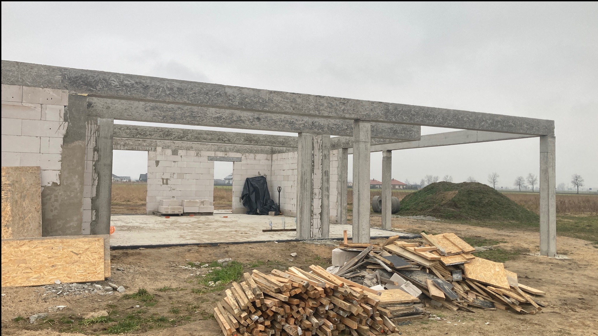 Surowy stan domu w budowie z betonu komórkowego, widoczne betonowe słupy i nadproża, składowane deski szalunkowe i płyty OSB na placu budowy.