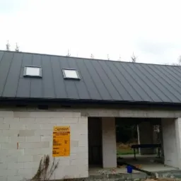 Szary dach z blachy na surowym budynku w Czerwińsku nad Wisłą, widoczne dwa okna dachowe i komin.