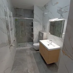 Nowoczesna łazienka po remoncie w Rzeszowie: kabina prysznicowa ze szklanymi drzwiami, umywalka z drewnianą szafką i lustro z oświetleniem, wykończona płytkami imitującymi marmur.