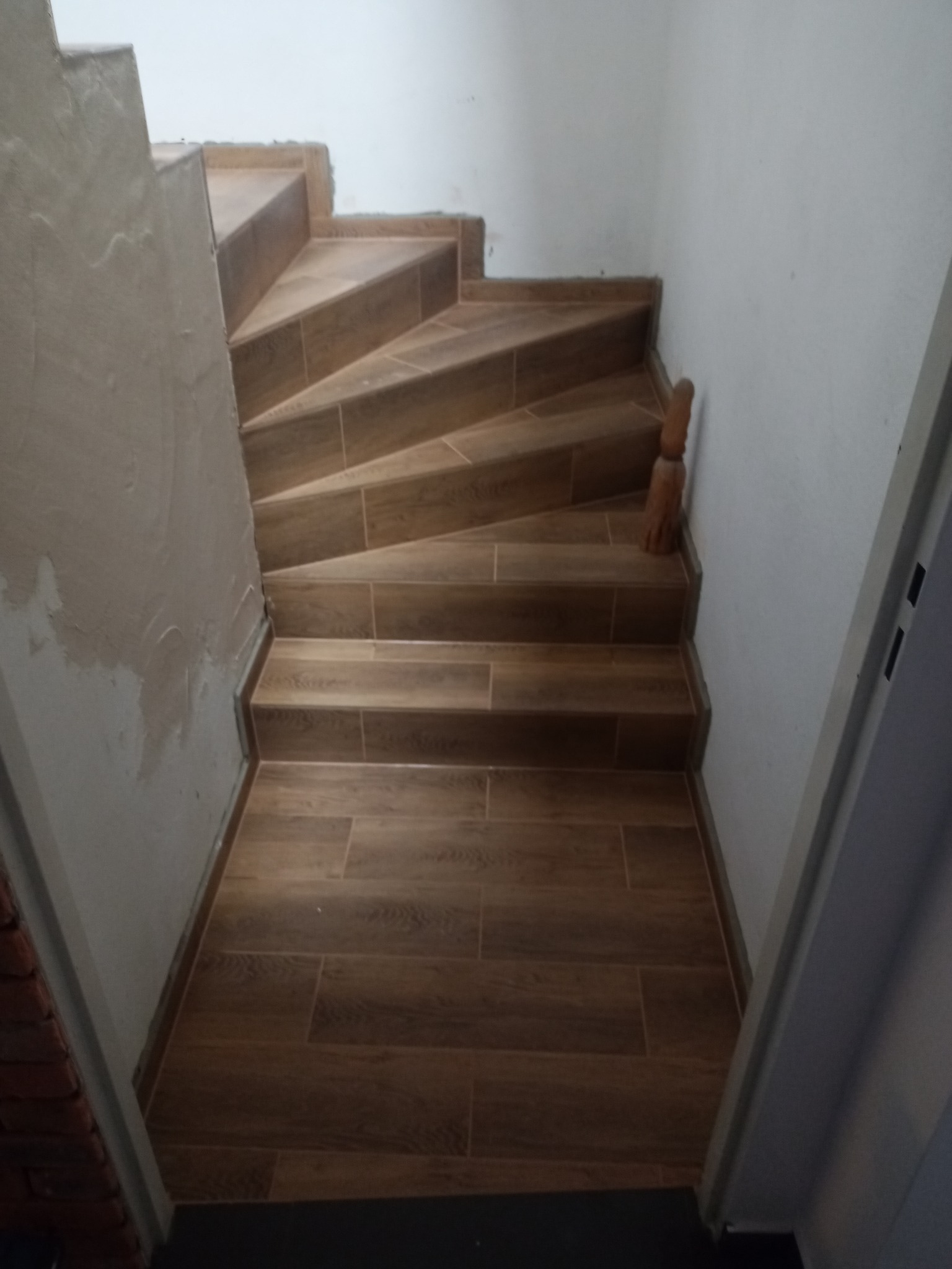 Wykończone schody wewnętrzne z płytek imitujących drewno, widok z góry, ściana po lewej częściowo otynkowana, ściana po prawej pomalowana na biało.