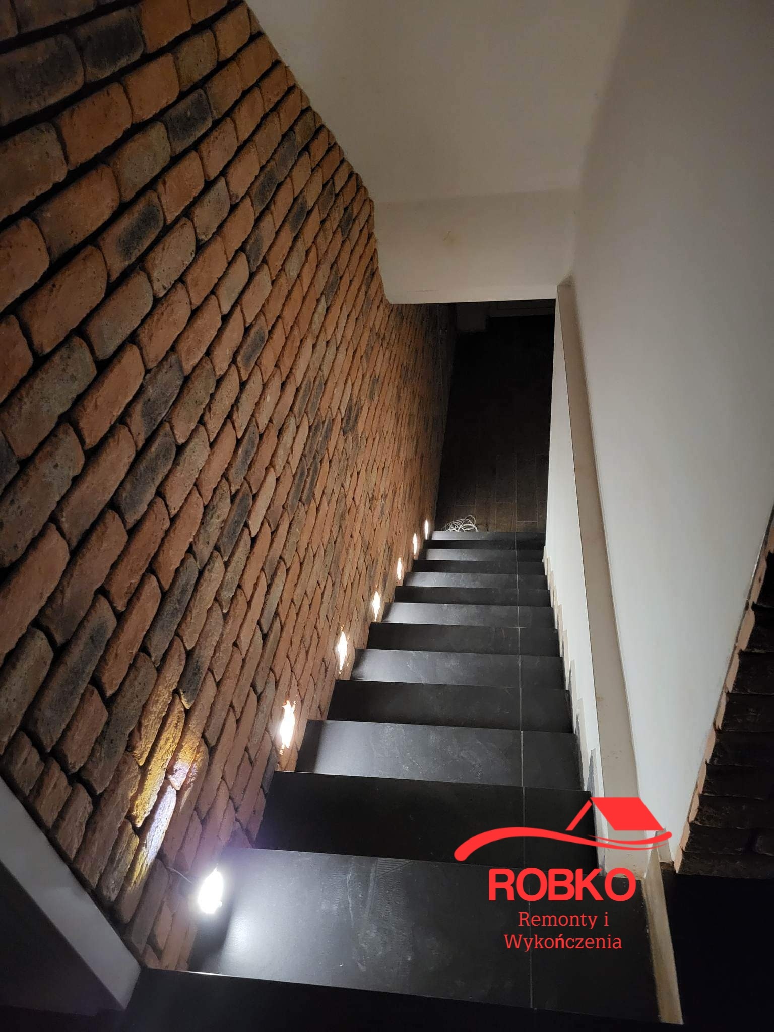 Schody z czarnymi stopniami, oświetleniem punktowym w ścianie z cegły klinkierowej i białą ścianą po prawej stronie, widoczne logo firmy ROBKO.