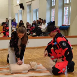 Uczennica w bluzce w kwiaty ćwiczy resuscytację krążeniowo-oddechową na fantomie pod nadzorem instruktora w czerwonym kombinezonie, w tle grupa uczniów w sali z lustrami.