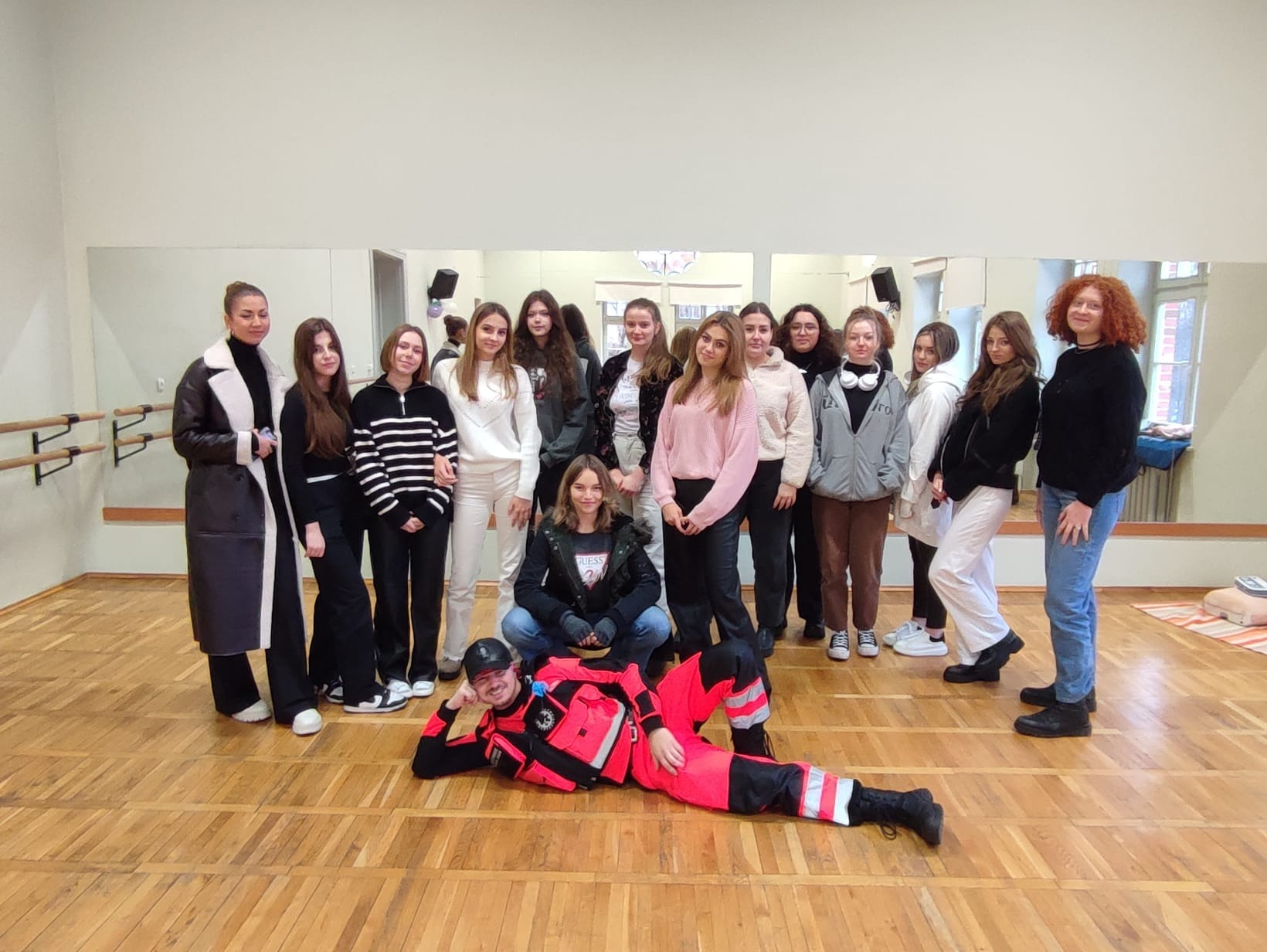 Grupa nastolatek i instruktor w stroju ratownika medycznego podczas kursu pierwszej pomocy; instruktor leży na podłodze, dziewczyny stoją za nim.