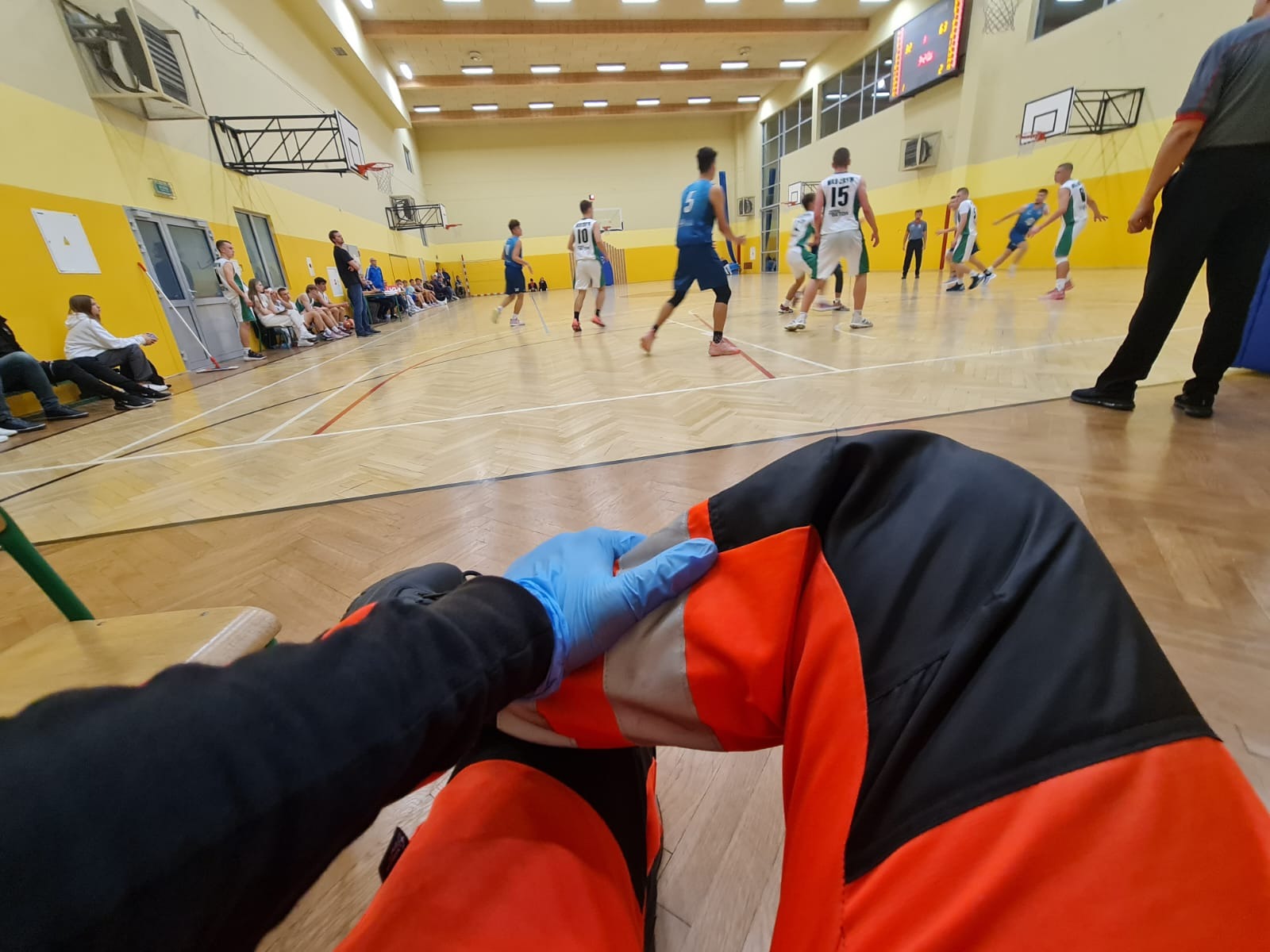 Medyk w pomarańczowo-czarnym ubraniu i niebieskiej rękawiczce siedzi na ławce, obserwując mecz koszykówki na hali sportowej.