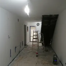 Wnętrze w trakcie odświeżania: zabezpieczona podłoga, ściany przygotowane do malowania, dwie drabiny, radio budowlane i napój na jednej z nich.