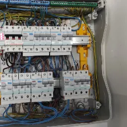 Otwarta skrzynka bezpiecznikowa z zainstalowanymi bezpiecznikami i okablowaniem, widoczne żółte elementy mocujące szyny DIN. Jeden z bezpieczników w pozycji 'ON' świeci na czerwono.