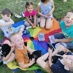 HULA HOP ANIMACJE - Grupa roześmianych dzieci siedzi na kolorowej chuście animacyjnej na trawie podczas zabawy na świeżym powietrzu.