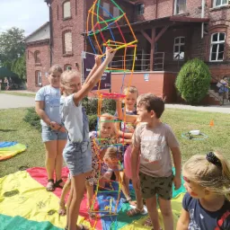 Grupa dzieci buduje wysoką konstrukcję z kolorowych rurek na tle ceglanego budynku podczas imprezy plenerowej, stojąc na kolorowej plandze.
