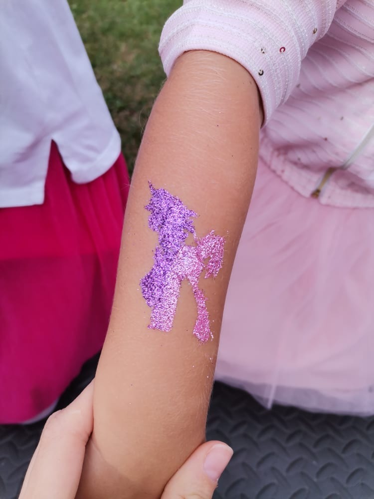 Brokatowy tatuaż jednorożca w odcieniach fioletu i różu na dziecięcym ramieniu, widoczne fragmenty różowej sukienki i białej bluzki dziecka.