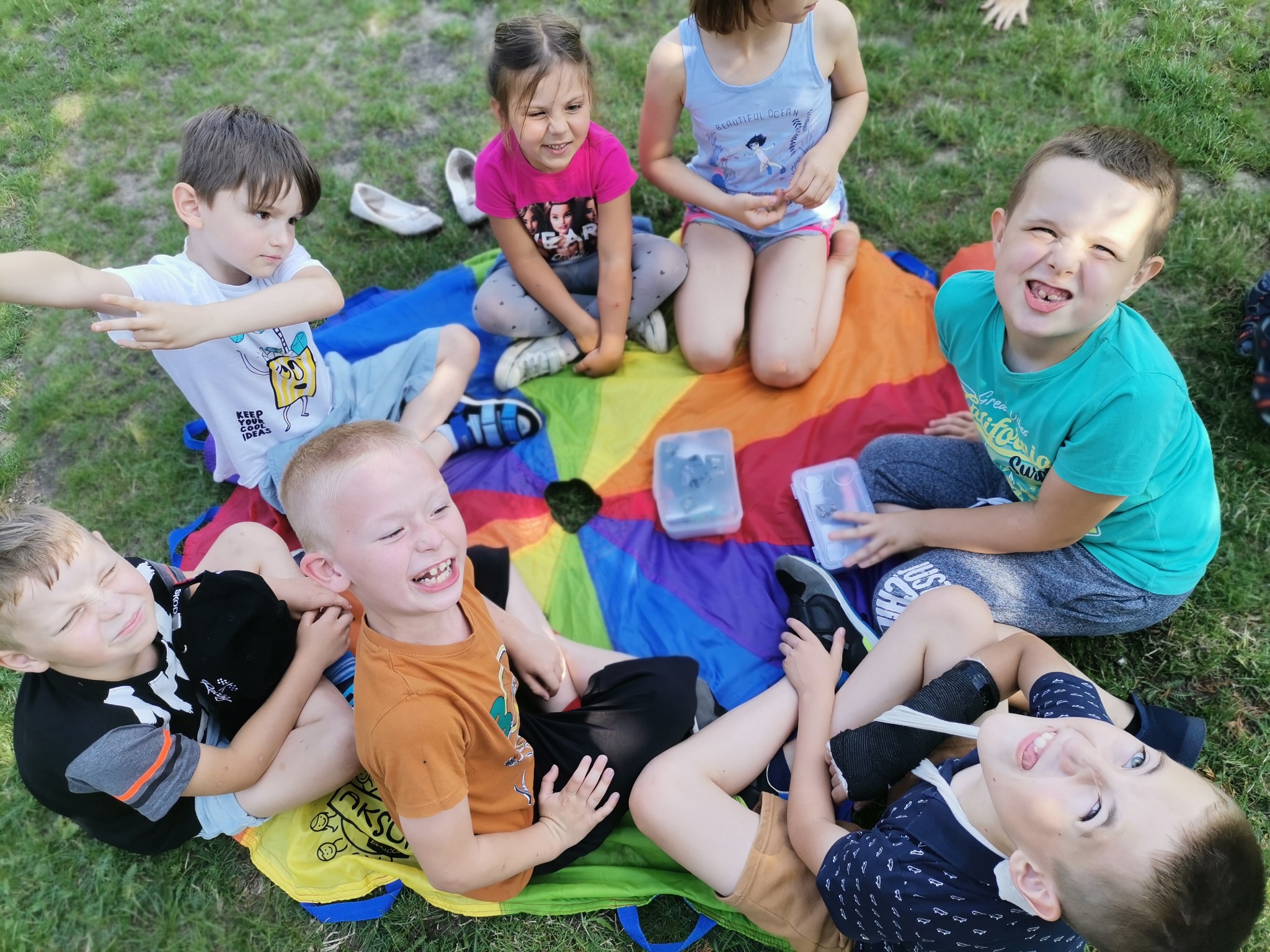 Grupa roześmianych dzieci siedzi na kolorowej chuście animacyjnej na trawie podczas zabawy na świeżym powietrzu.