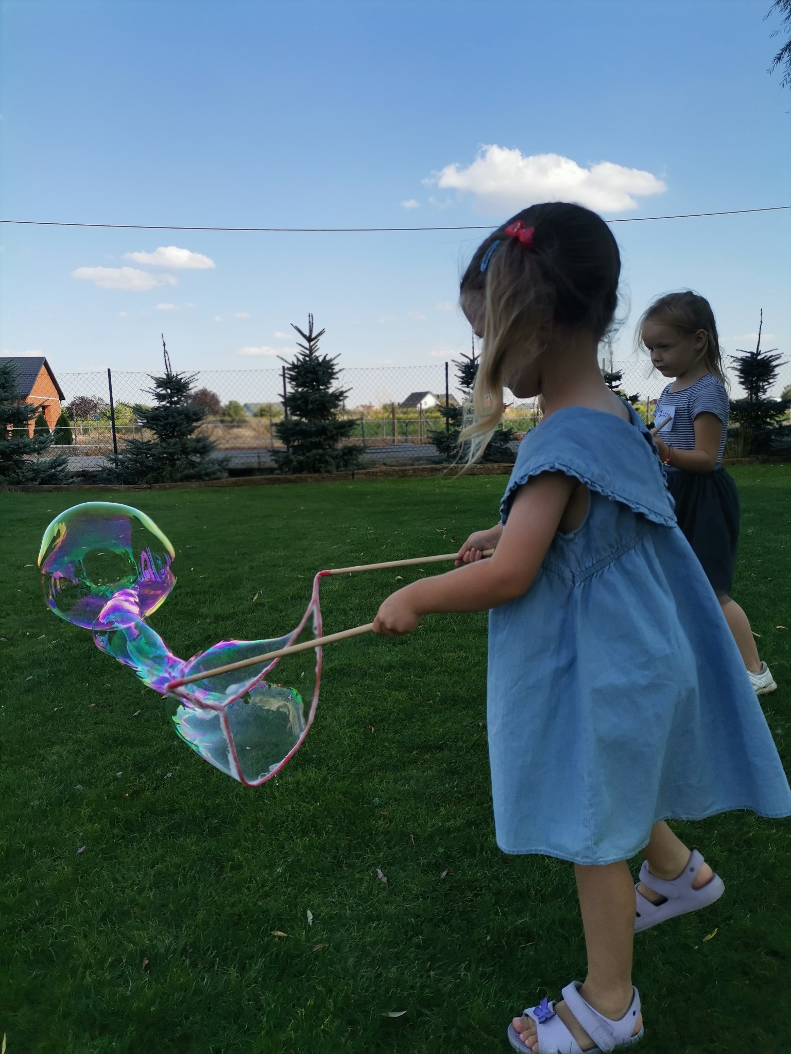 Dziewczynka w dżinsowej sukience puszcza ogromne, opalizujące bańki mydlane na trawniku, druga dziewczynka obserwuje w tle.