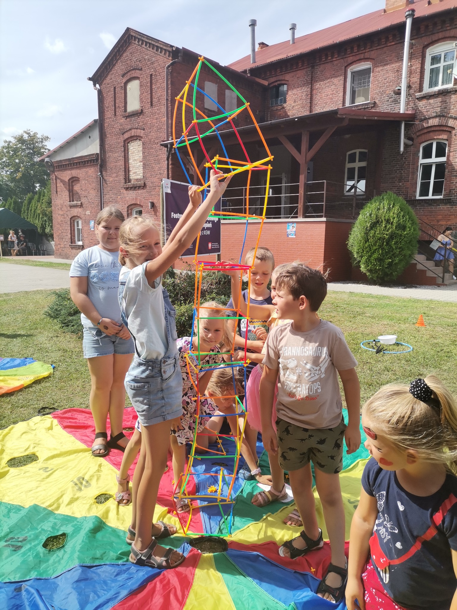 Grupa dzieci buduje wysoką konstrukcję z kolorowych rurek na tle ceglanego budynku podczas imprezy plenerowej, stojąc na kolorowej plandze.